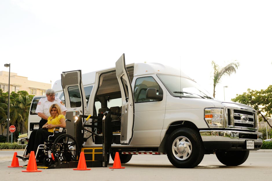 A white wheelchair-accessible van parked on a quiet street with a wide side door open, revealing a woman in a yellow top seated in a wheelchair being assisted by two men during a home relocation proce