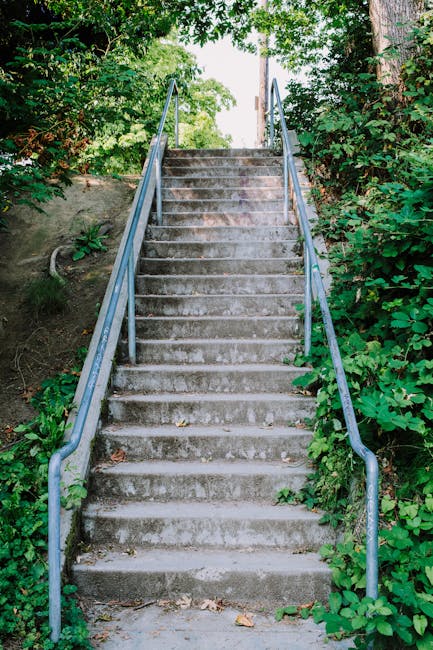 A wide concrete staircase inside a building, with metal handrails on both sides and in the middle, leading upwards. The staircase is flanked by a red brick wall on the left and a roughened concrete wa