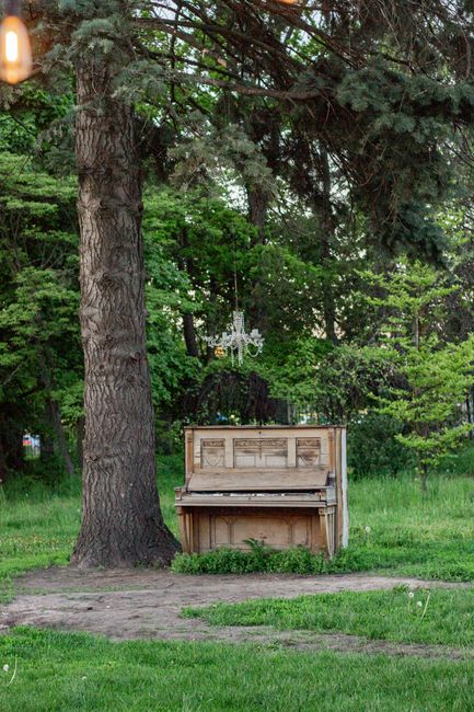 A vintage upright piano with a worn black finish, positioned outdoors against a vertical wooden fence made of narrow, evenly spaced light brown planks. The piano's front panel is open, exposing the in